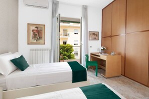 A hotel room with a bed, a desk, a wardrobe, and a balcony with a view of buildings and greenery.