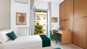 Habitación de hotel con cama, escritorio, armario y balcón con vistas a edificios y vegetación.