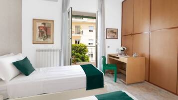 A hotel room with a bed, a desk, a wardrobe, and a balcony with a view of buildings and greenery.