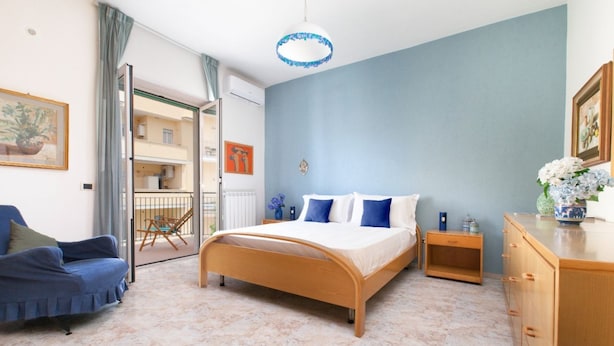 Un dormitorio con cama, cómoda de madera, silla azul y vista al balcón exterior.