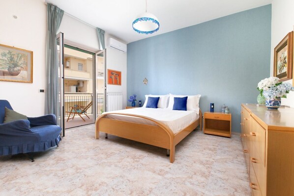 A bedroom with a bed, a wooden dresser, a blue chair, and a view of the outside balcony.