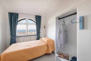 A bedroom with a bed, a window with curtains, a wardrobe with clothes, and a view of the sea.