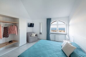 Une chambre à coucher avec un lit, un fauteuil gris, une télévision, une fenêtre avec des rideaux et une armoire remplie de vêtements.