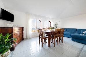 A dining area with a wooden table and chairs, a blue sofa, a TV mounted on the wall, and a balcony with a view.