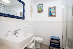 Un baño con lavamanos, inodoro y ducha blancos. Hay tres cuadros colgados en la pared y un atrapasueños.