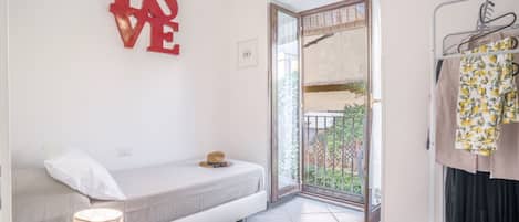 Una habitación pequeña y moderna con cama, mesita de noche con lámpara, decoración con la palabra 'LOVE' en rojo, perchero y puerta al balcón.