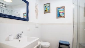 Un baño con lavamanos, inodoro y ducha blancos. Hay tres cuadros colgados en la pared y un atrapasueños.