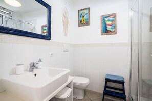 Een badkamer met een wit wastafelmeubel, toilet en douche. Er hangen drie schilderijen en een oogster. 