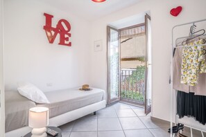 Een kleine, moderne kamer met een bed, een nachtkastje met een lamp, een rode 'LOVE' decoratie, een kledingrek en een openslaande balkondeur.