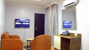 A hotel room with a TV mounted on the wall, a desk with a coffee maker, and two orange chairs. - Caritas Inn Ilasan (Lekki)