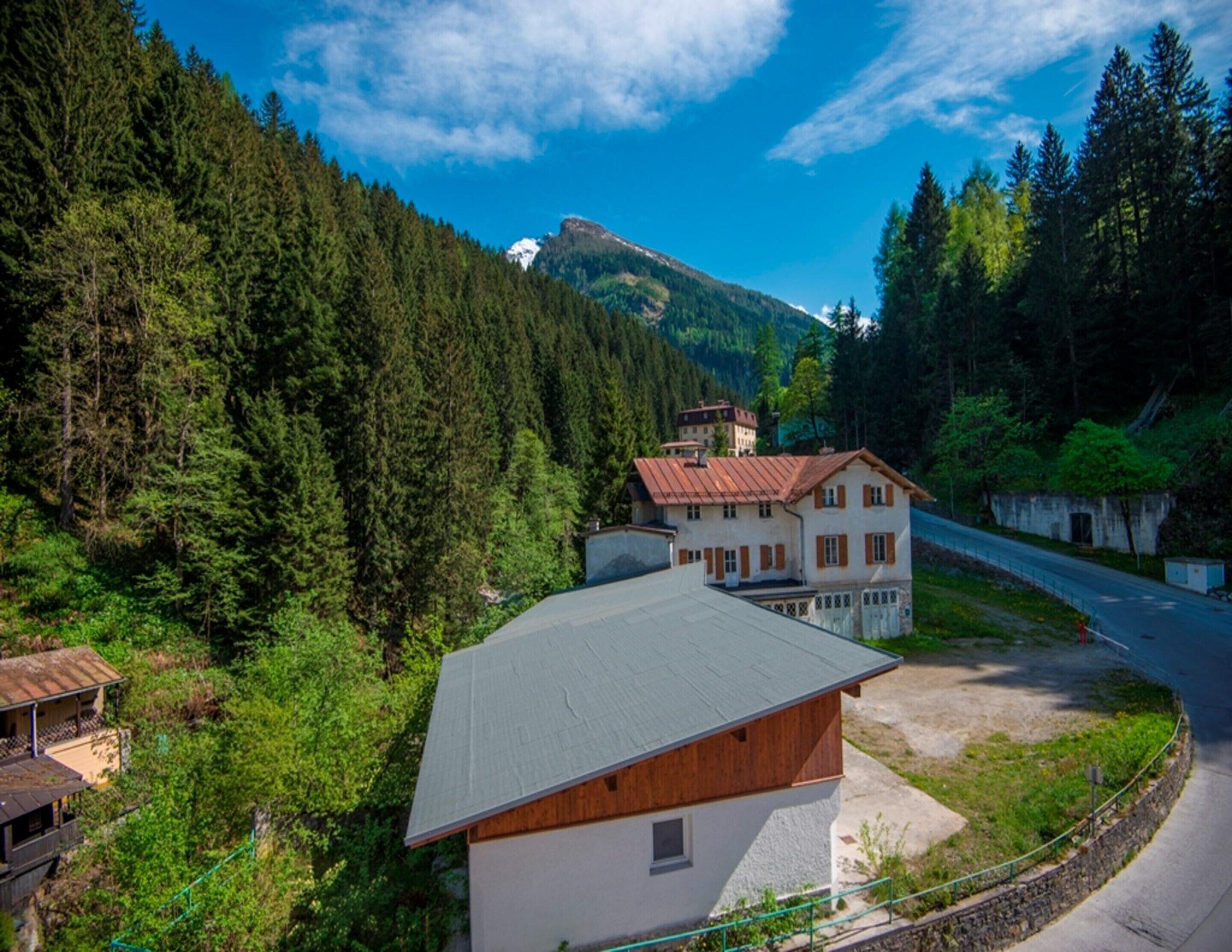 A two-story building with a sloped roof, surrounded by dense forest and a mountain in the background.