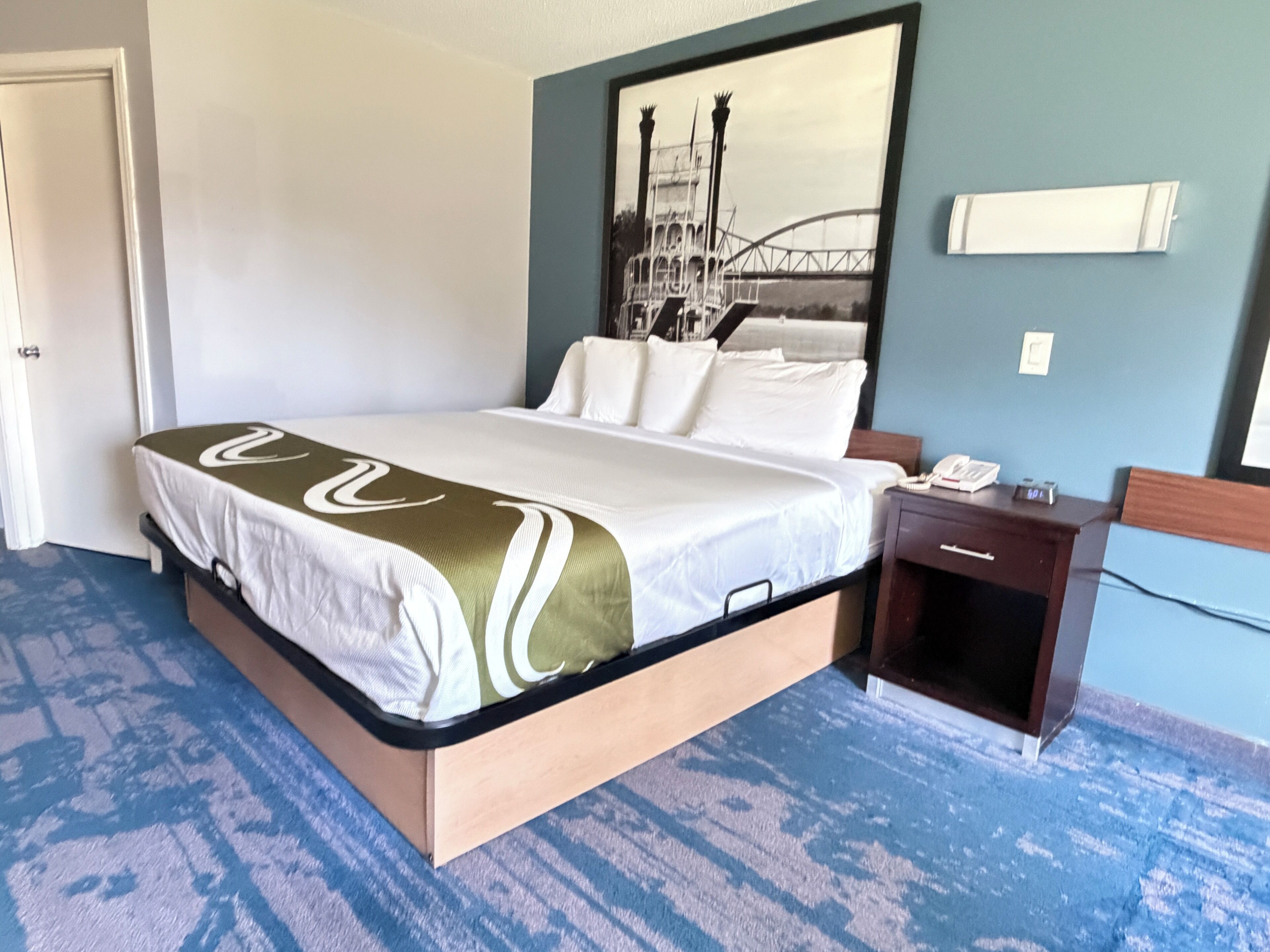 Quarto de hotel com cama, mesa de cabeceira, telefone e luminária fixada na parede.