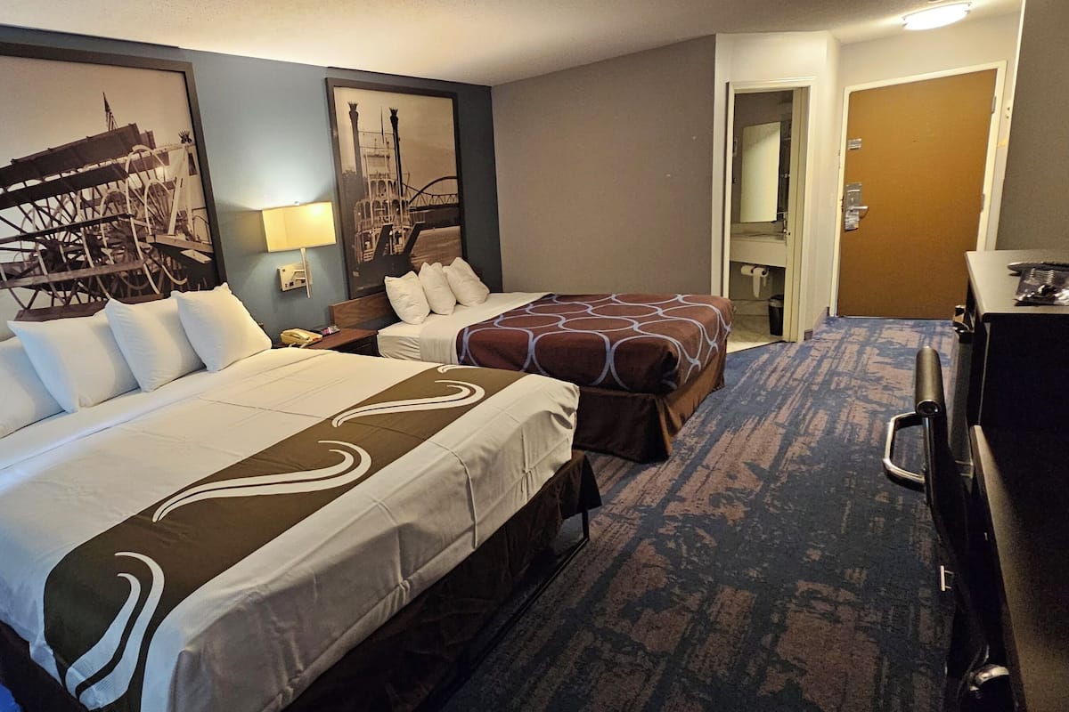 A hotel room with two beds, a desk, a chair, and a bathroom visible through an open door.
