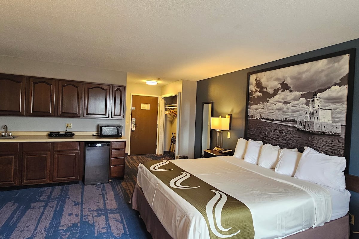 A hotel room with a bed, a desk with a microwave, a small kitchenette, and a large painting on the wall.