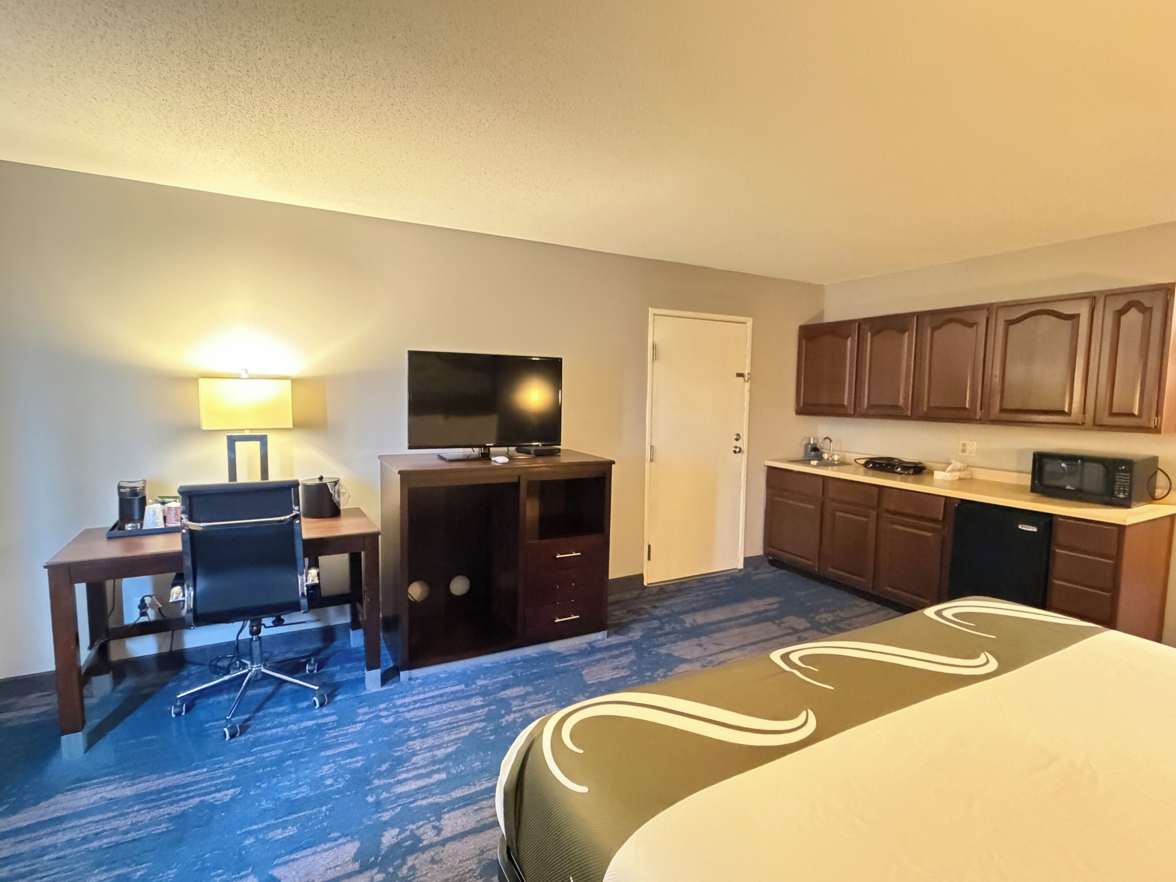 A hotel room with a bed, desk, chair, television, kitchenette, and a door.