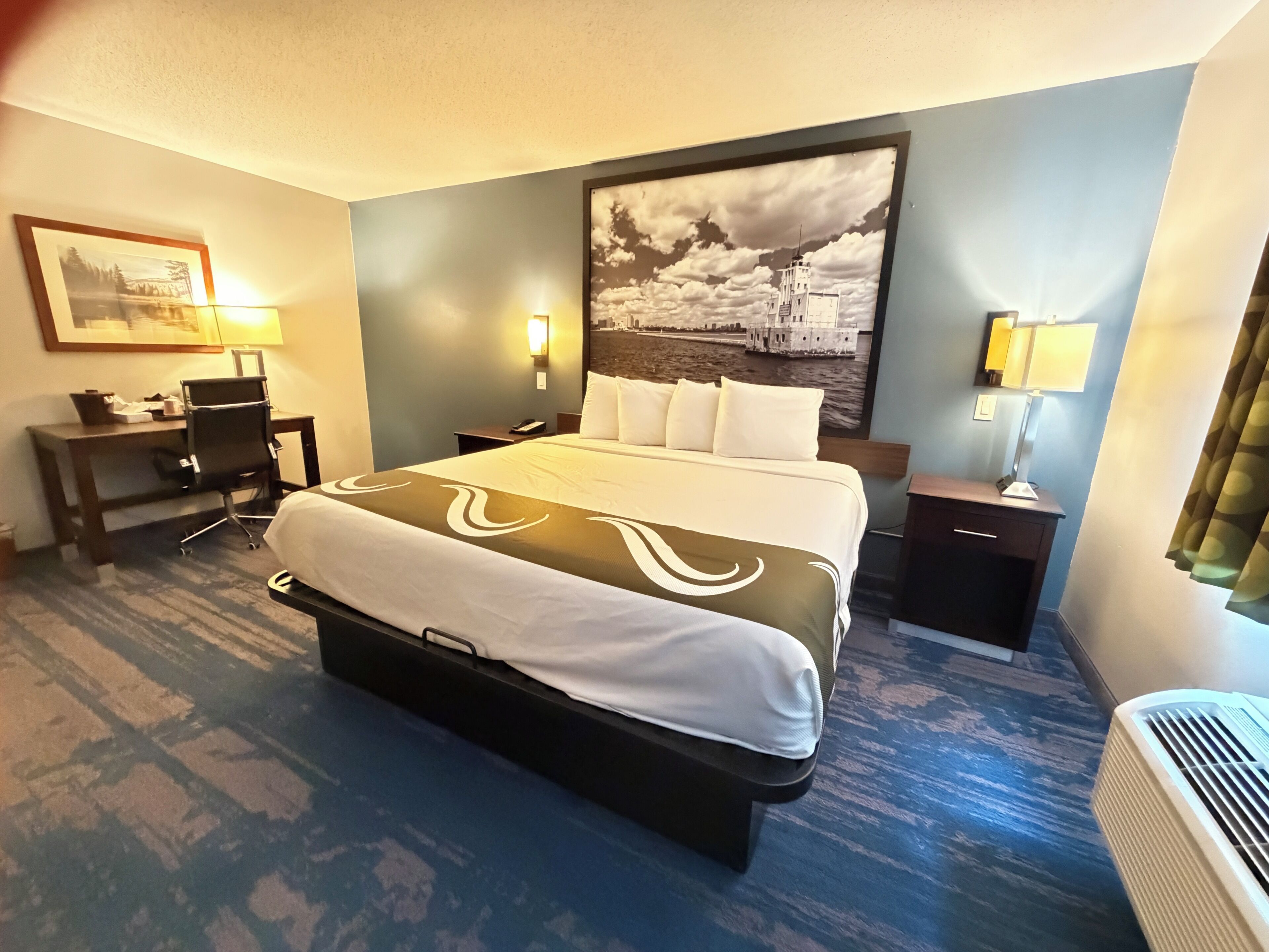 A hotel room with a bed, desk, chair, lamps, and a large wall painting.