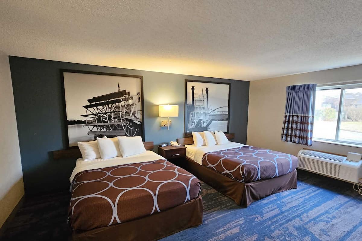 A hotel room with two beds, a large window with curtains, a wall-mounted lamp, and a radiator.