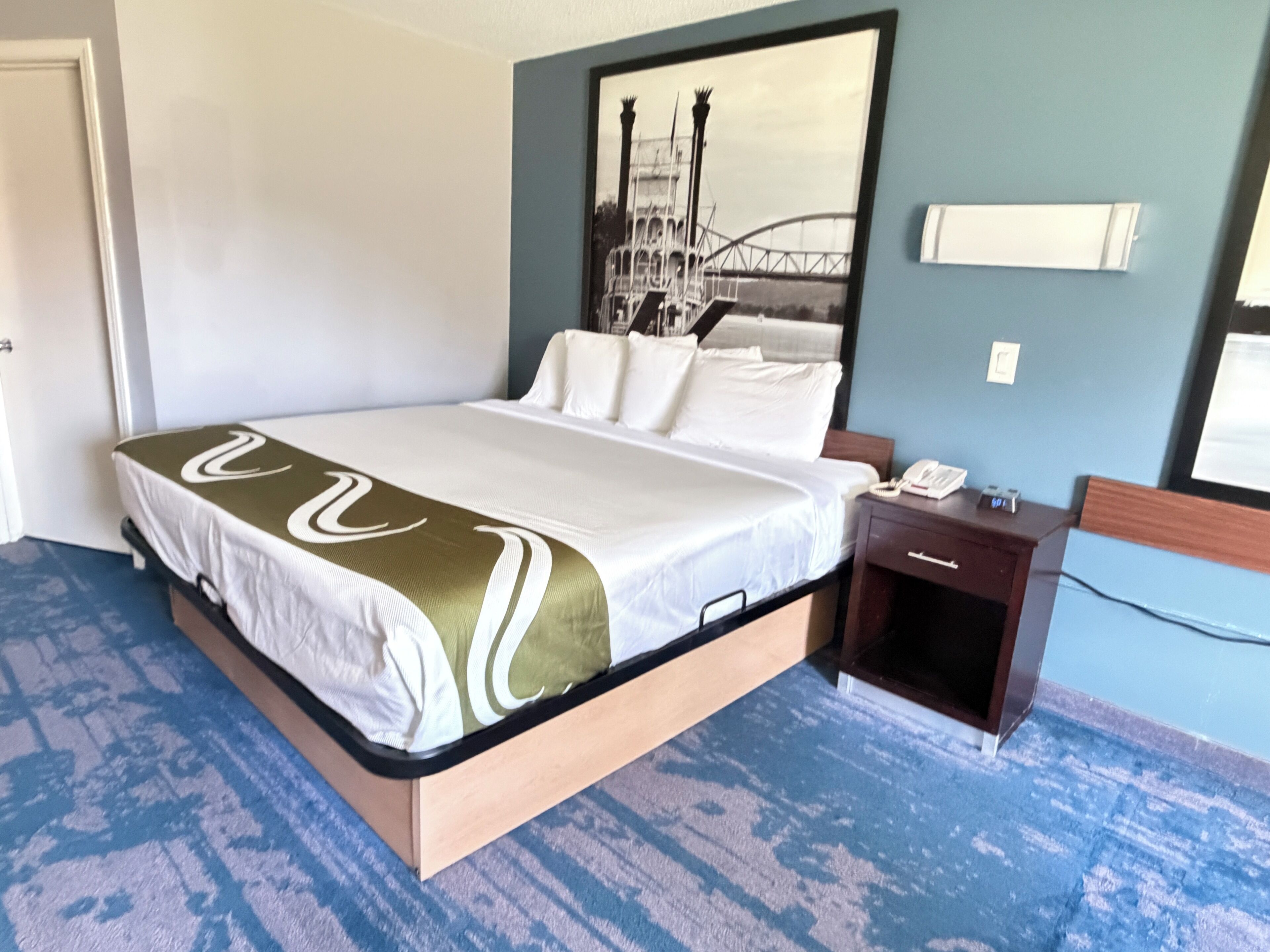 A hotel room with a bed, a nightstand, a framed picture on the wall, and a carpeted floor.