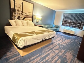 A hotel room with a large bed, a sofa, a window with curtains, and a wall-mounted air conditioner. - Northwood Inn Prairie Du Chien (Prairie Du Chien)
