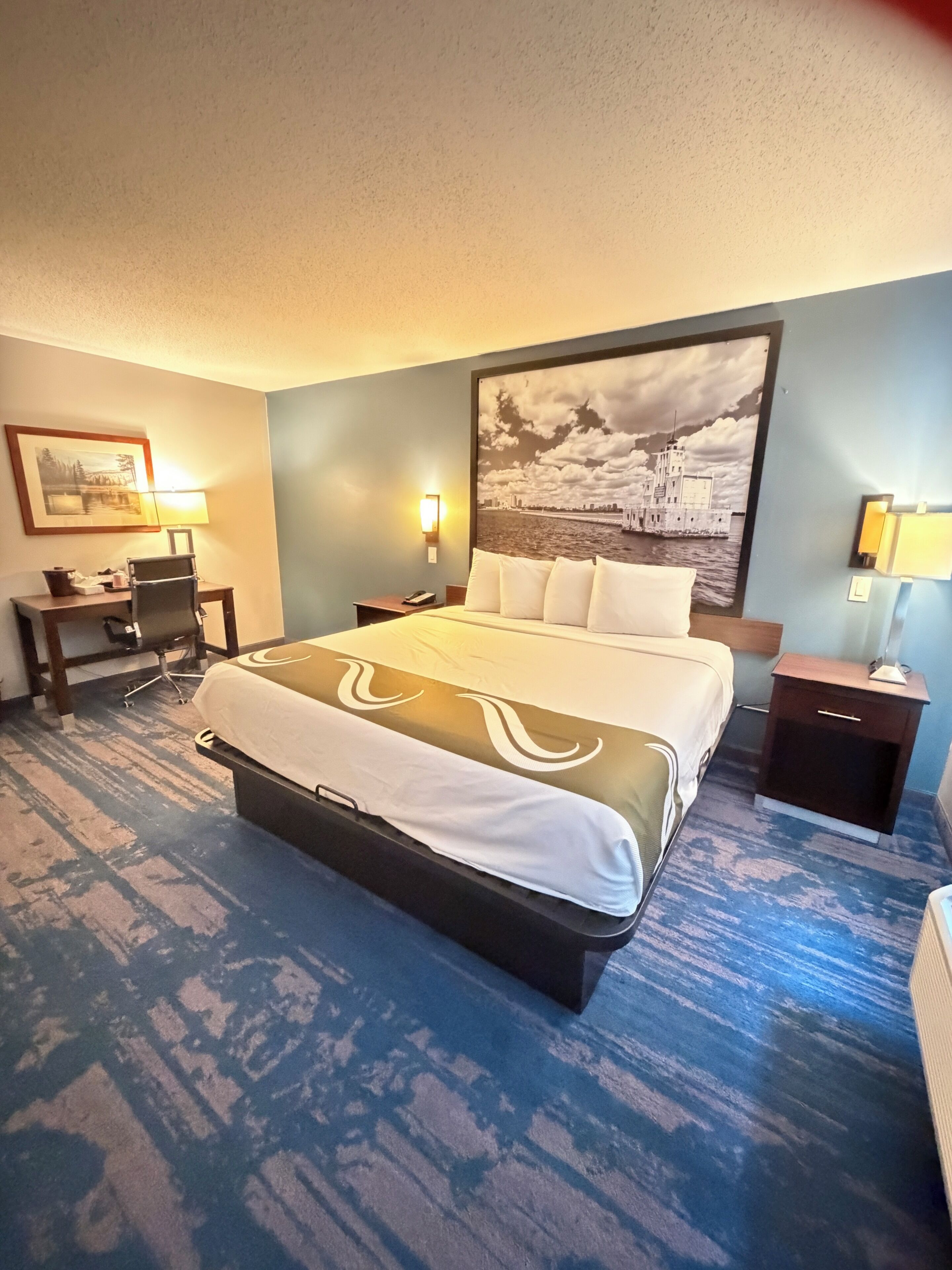 Une chambre d’hôtel avec un grand lit, un bureau, une chaise, une table de chevet et une lampe fixée au mur.