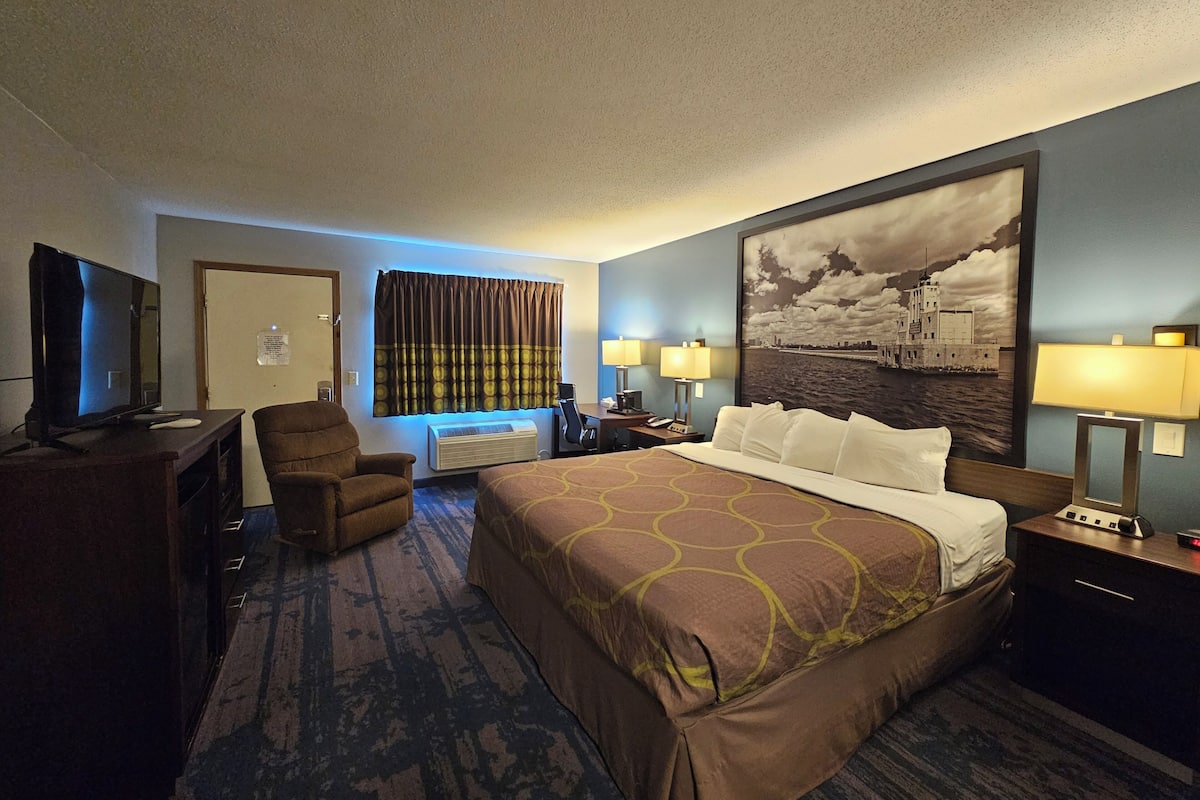 A hotel room with a large bed, a desk with a TV, a chair, a window with curtains, and a painting on the wall.
