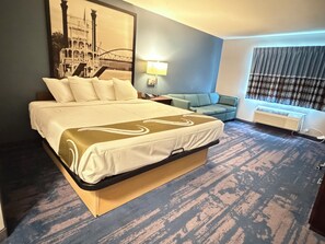 Quarto de hotel com uma cama grande, um sofá, uma luminária e uma janela com cortinas.