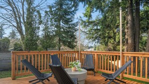 Eine Holzterrasse mit einem runden Tisch und zwei Stühlen, umgeben von einem Hollerleger und Bäumen.