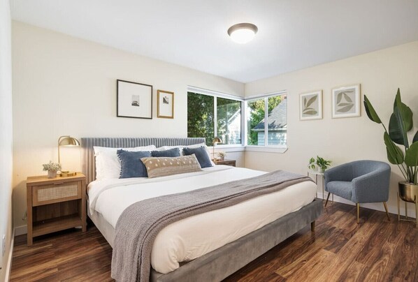A bedroom with a large bed, a nightstand, a chair, and a window with a view of greenery.