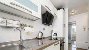 Une cuisine moderne équipée d’un évier en acier inoxydable, d’une bouilloire, d’une machine à café et d’un four à micro-ondes. Les meubles sont blancs et le plan de travail est surmonté d’un pare-feu brun.