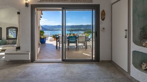 Une pièce dotée d’une porte-fenêtre coulissante donnant sur une terrasse extérieure avec une table et des chaises, un pot de fleurs, et une vue sur la mer et les collines.