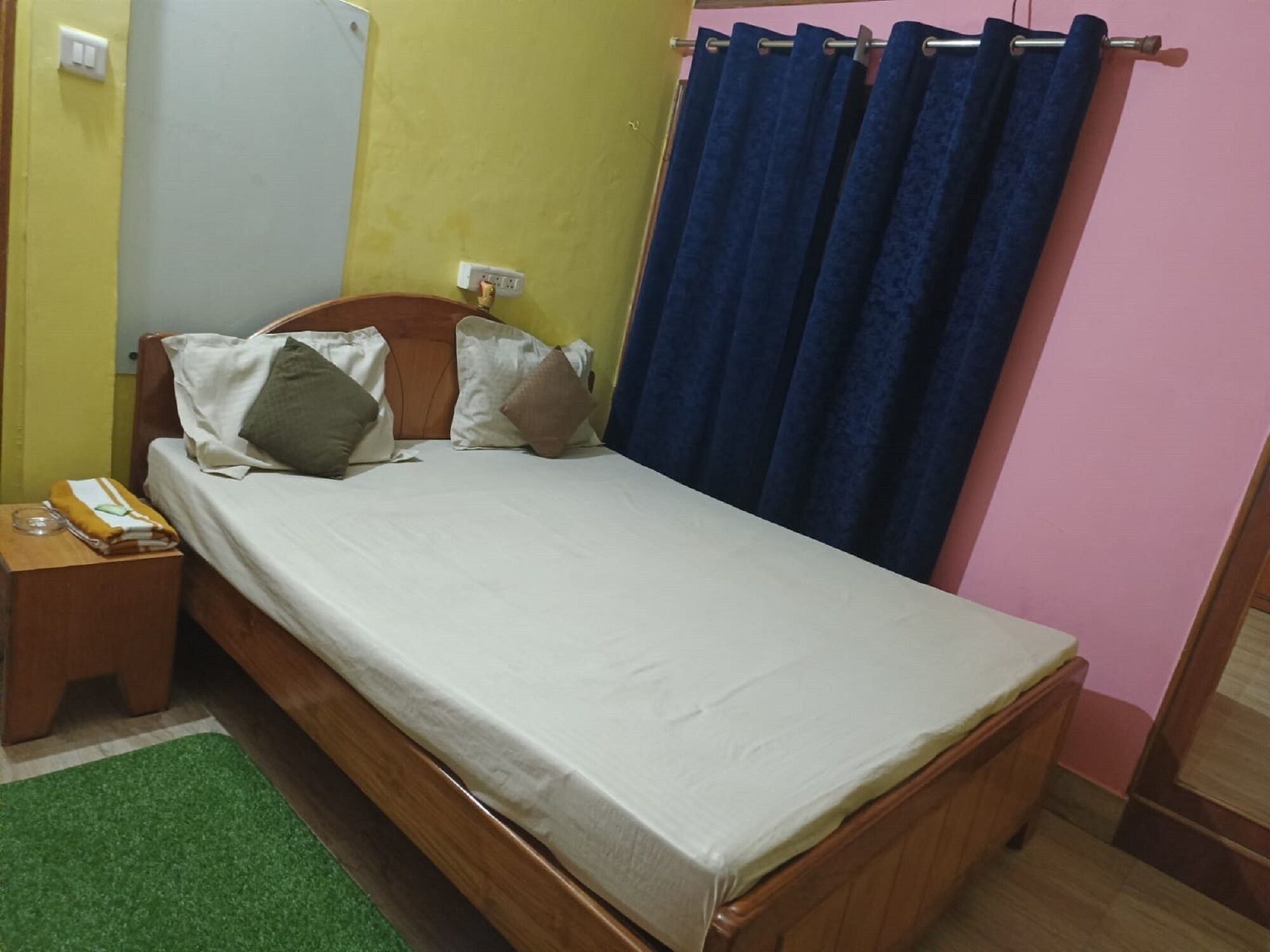 A single bed with a wooden frame, a green rug, and a bedside table with a lamp.