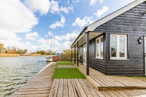 A wooden dock leading to a dark-colored cabin with a covered porch and a small grassy area. - Host Stay Tide Time (Great Yarmouth)