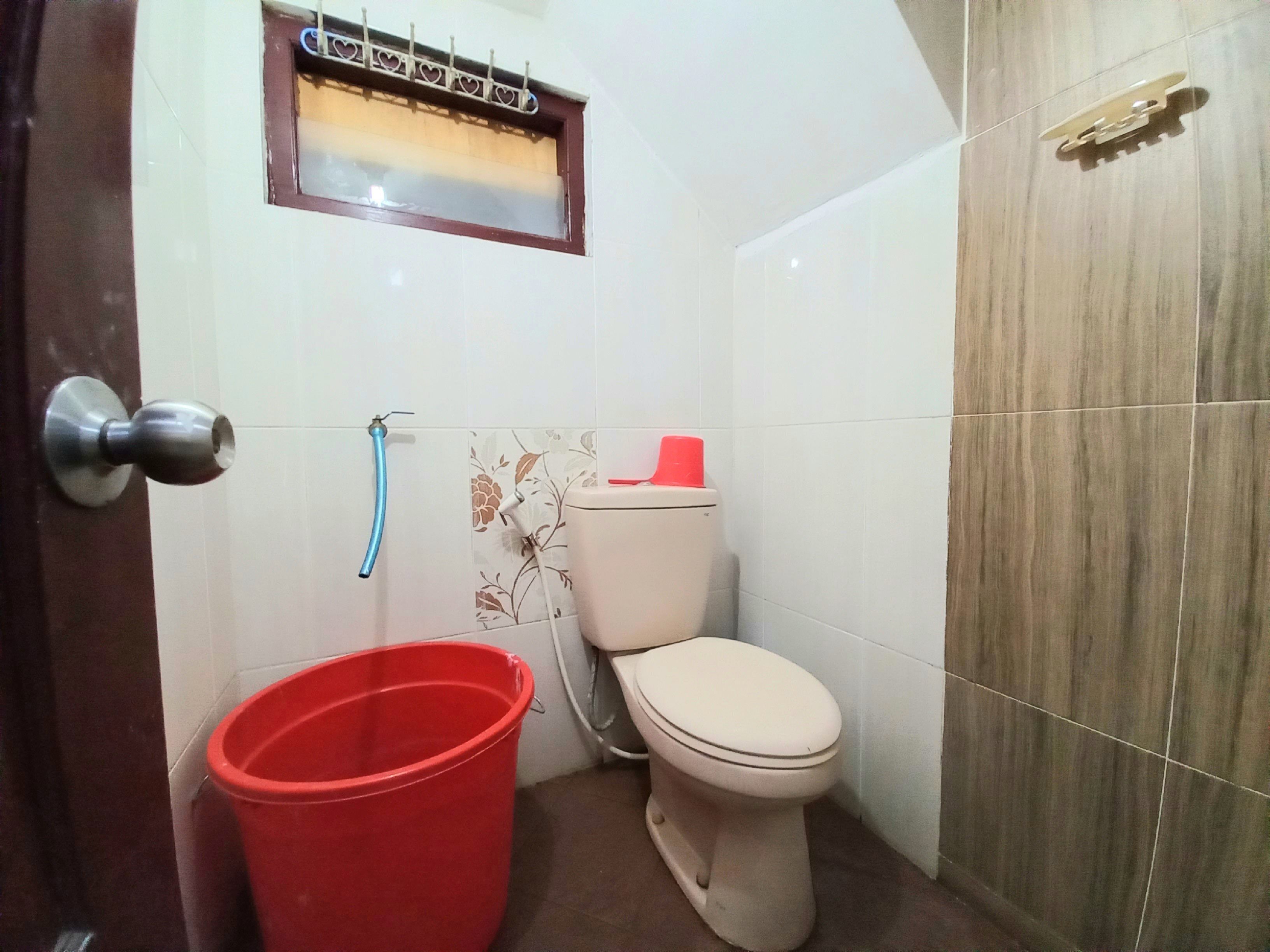 Un baño pequeño con inodoro, un cubo rojo, una manguera azul y una ventana con diseño de corazón.