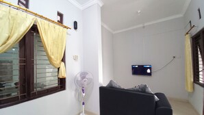 Un soggiorno con un divano, una ventola a piantana, una TV fissata al muro e una finestra con tende.