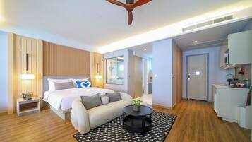 A hotel room with a bed, a sofa, a small table with a plant, a ceiling fan, and a kitchenette.