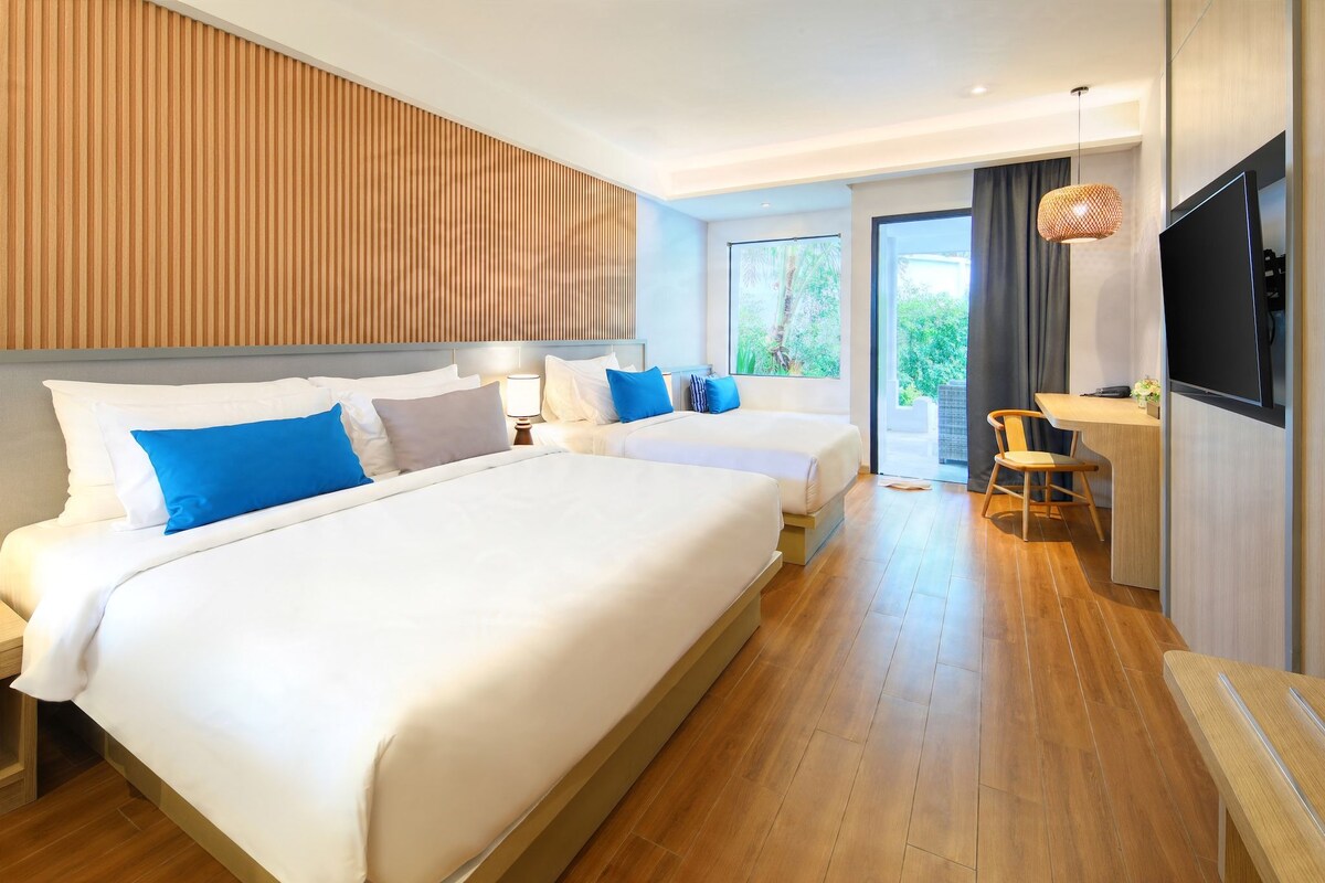 A hotel room with two beds, a desk, a TV, and a view of greenery outside.