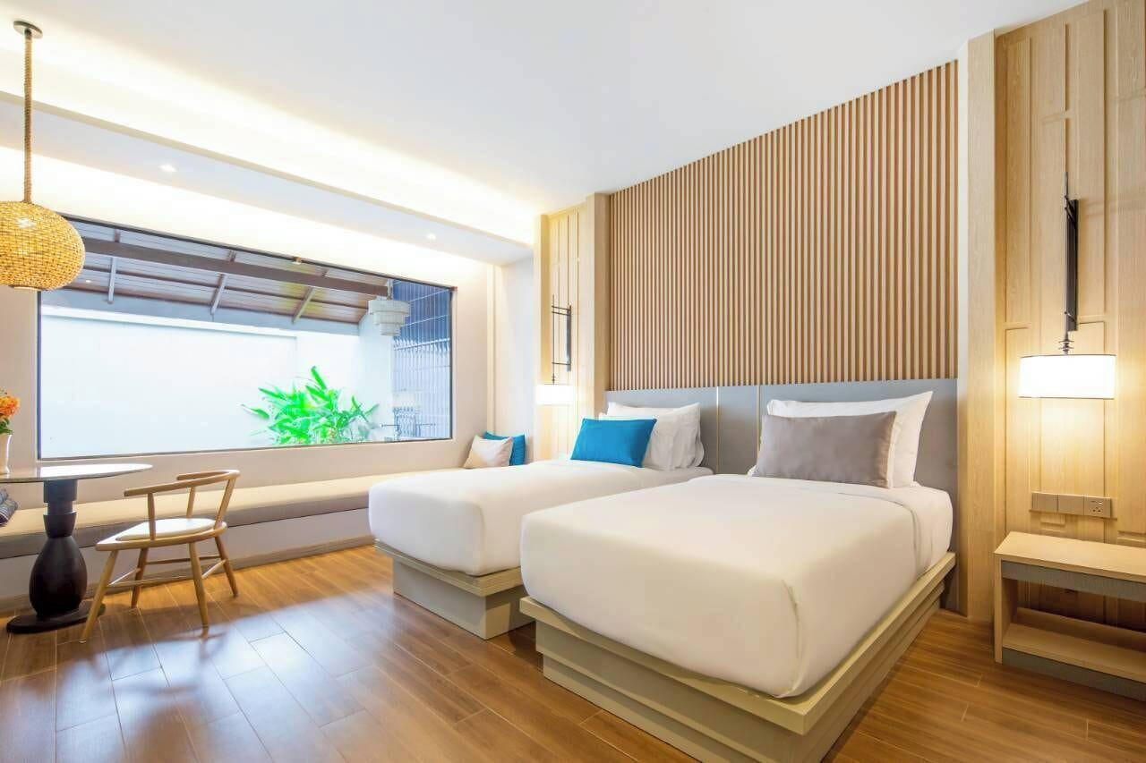 A modern hotel room with two beds, a large window with a view of greenery, a dining area with a table and chairs, and a wooden accent wall.