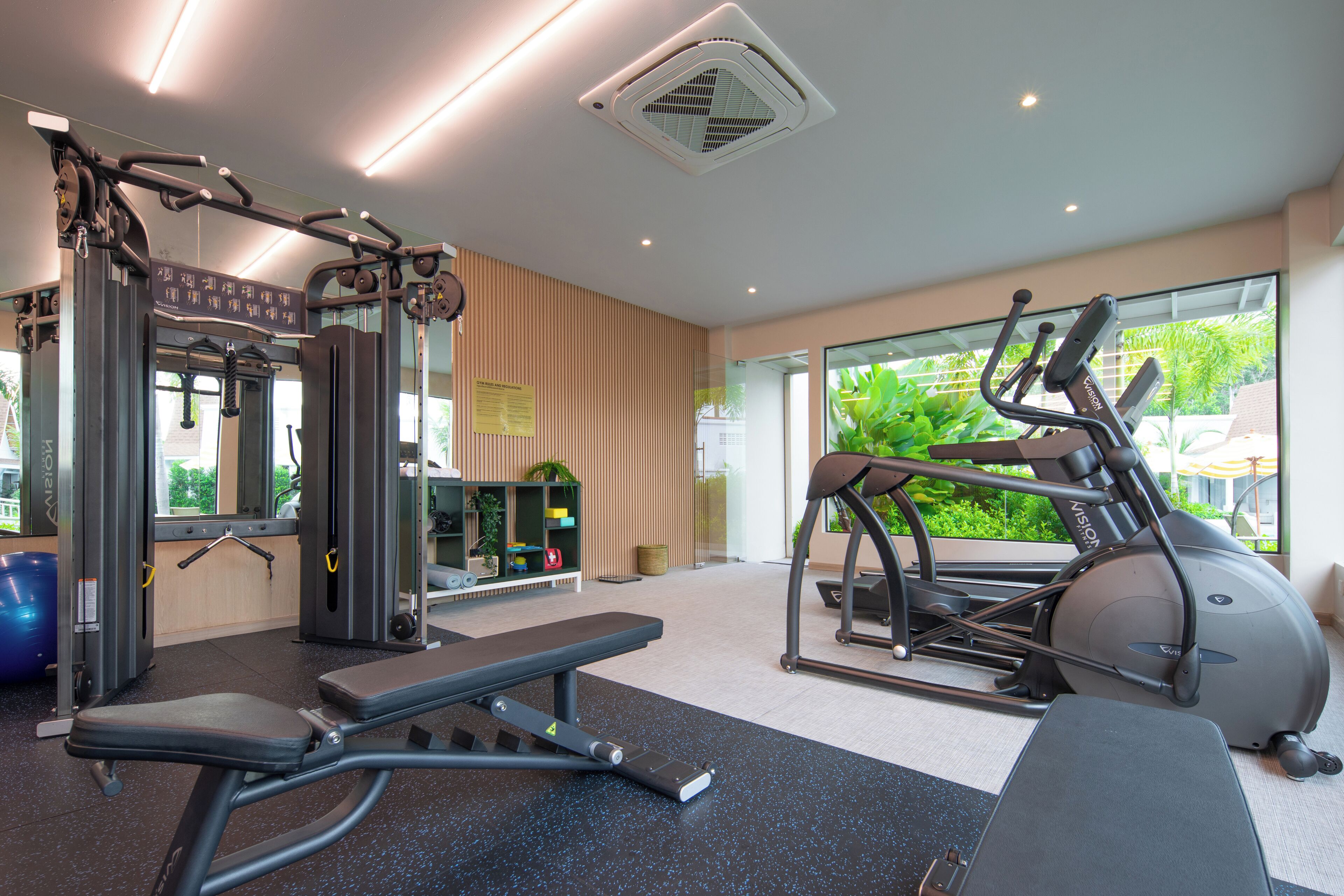Un gimnasio con equipo de ejercicio, una banca y un ventanal con vistas a zonas verdes.