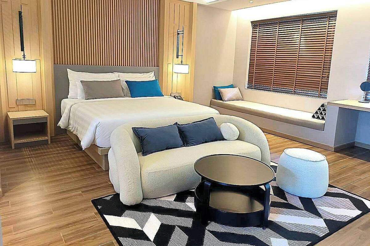 Een moderne hotelkamer met een groot bed, nachtkastjes, een bank, een ronde salontafel en een raam met jaloezieën.