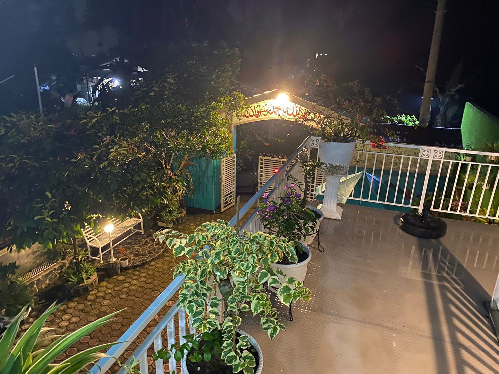 Une vue nocturne d’une cour intérieure avec un escalier, des plantes en pot et une arche décorative.