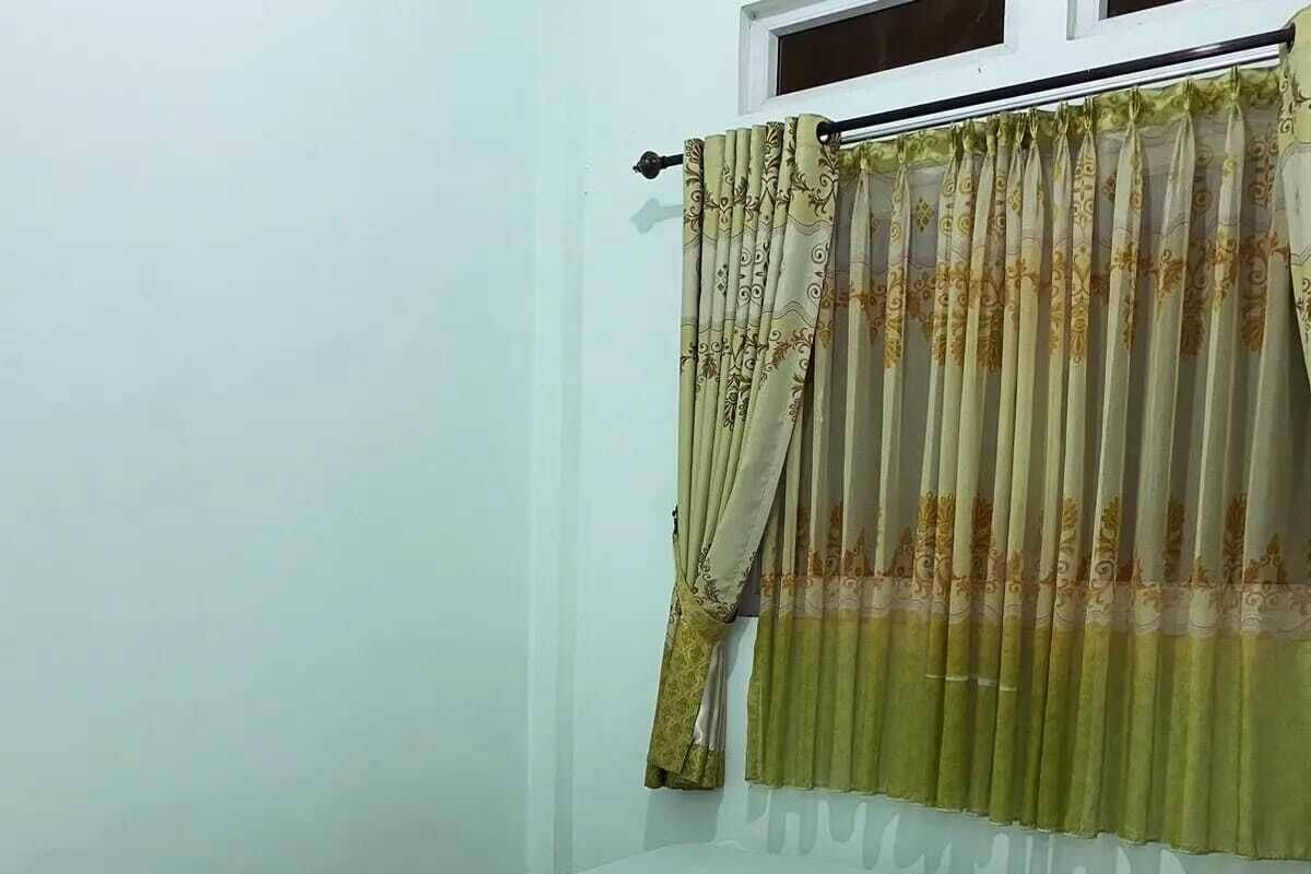 Ein kleines Zimmer mit einem Einzelbett, einer schlichten weißen Matratze, einem Fenster mit Vorhang und einer schlichten weißen Wand.