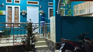 Une façade bleue avec une moto garée sur le côté droit, un pot de fleurs au premier plan et un escalier menant à une porte.