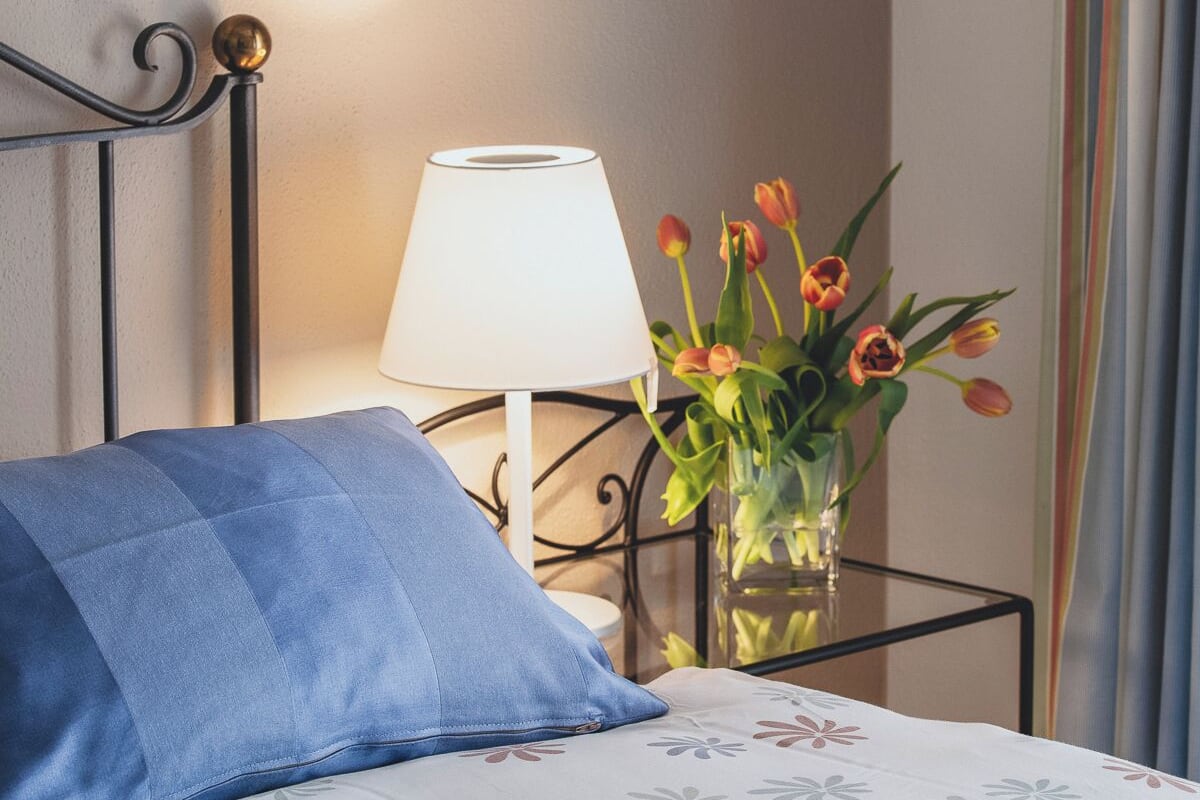 A neatly made bed with a floral-patterned bedspread, a blue pillow, a white bedside lamp, and a vase with tulips on a bedside table.