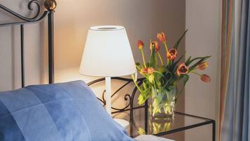 A neatly made bed with a floral-patterned bedspread, a blue pillow, a white bedside lamp, and a vase with tulips on a bedside table.