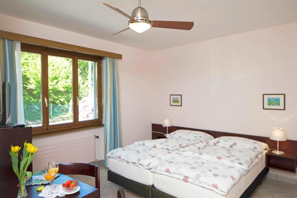 Ein Schlafzimmer mit einem Bett, zwei Nachttischen, einem Schreibtisch mit Stuhl, einem Fenster mit Vorhängen und einem Deckenventilator.