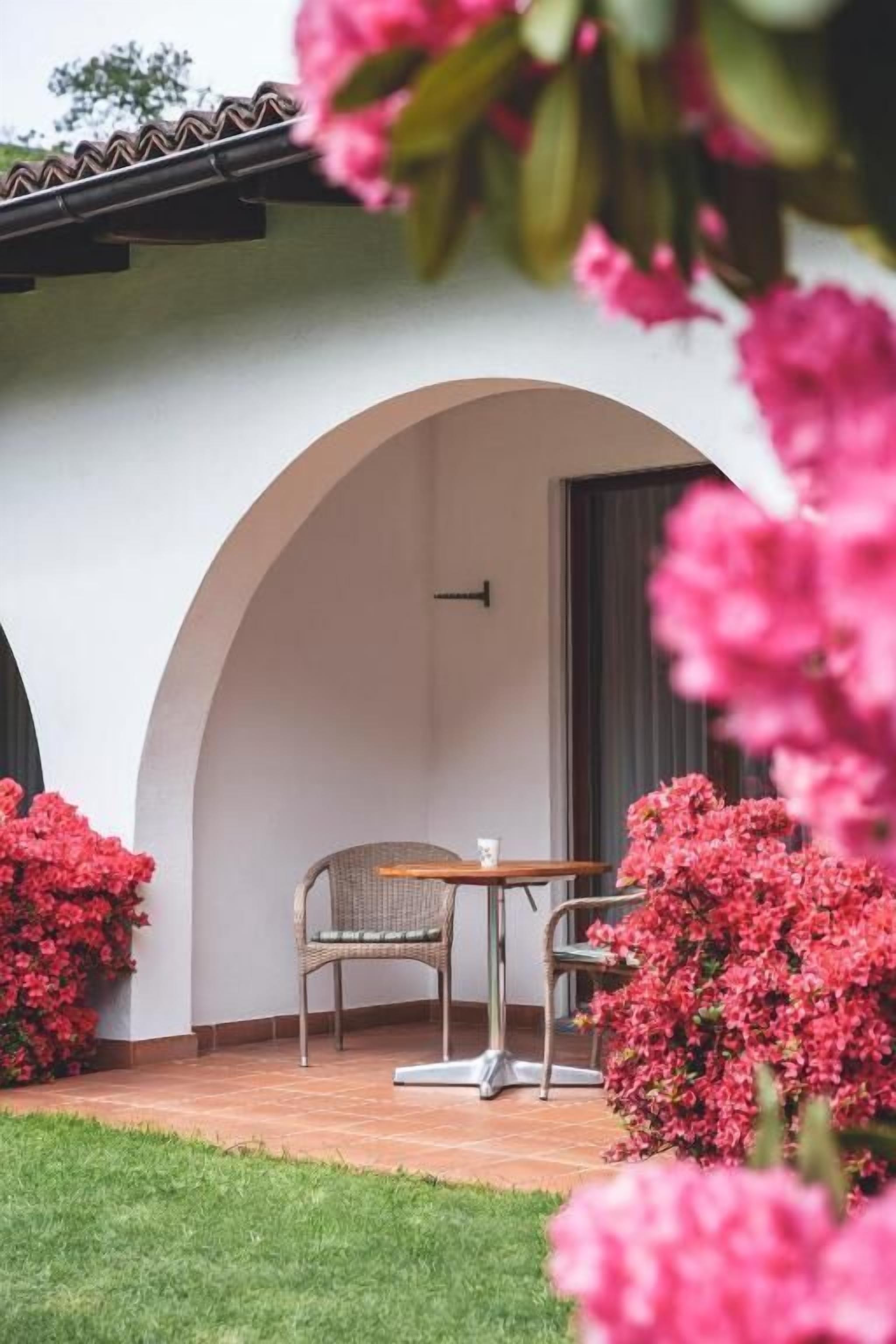 An outdoor patio with a table and chairs, surrounded by lush greenery and flowering plants.