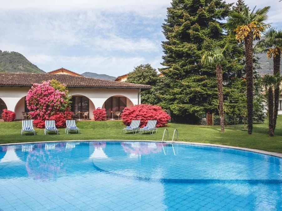 A swimming pool with lounge chairs, surrounded by lush greenery and palm trees, with a building in the background.