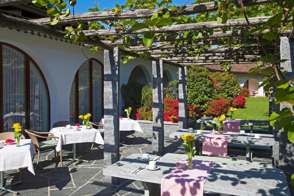 Un área de comedor al aire libre con mesas y sillas de piedra, cubierta por una pérgola de madera, rodeada de exuberante vegetación y flores.