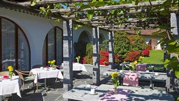 Un área de comedor al aire libre con mesas y sillas de piedra, cubierta por una pérgola de madera, rodeada de exuberante vegetación y flores.