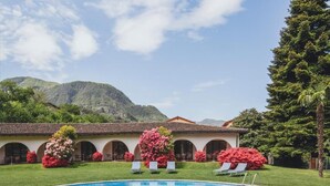 A swimming pool with lounge chairs, surrounded by lush greenery and a building with arched windows. - Garni Elena (Losone)